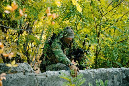 Man in uniform, climbs over the wall with sniper rifle on green forest background.