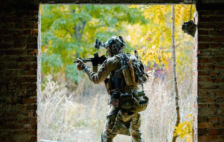 Man in uniform with machine gun between two walls on forest background. Airsoft soldier aims at the sight.の写真素材