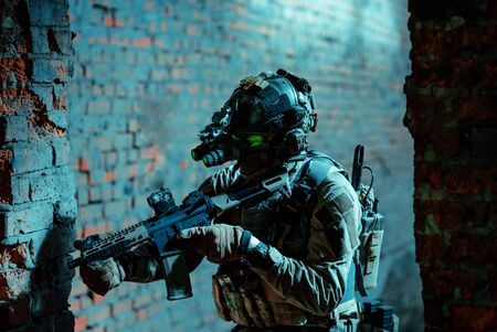 Man in uniform with machine gun and turned on night vision device move between brick wall. Closeup airsoft soldier with green light on face in night building.の写真素材