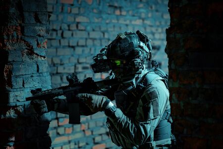 Man in uniform with machine gun and turned on night vision device move between brick wall. Closeup airsoft soldier with green light on face in night building.の写真素材