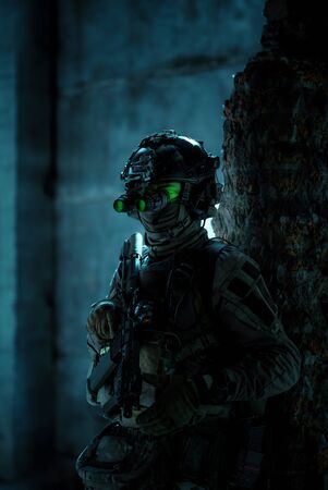 Man in uniform with machine gun and turned on night vision device move between brick wall. Airsoft soldier with green light on face in night building. Side view.の写真素材