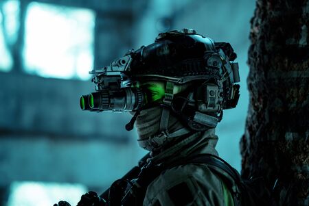 Closeup man uniform with machine gun and turned on night vision device. Airsoft soldier with green light on face in night building. Side view.