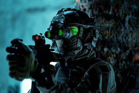 Man in uniform with machine gun and turned on night vision device beside brick wall. Closeup airsoft soldier with green light on face in night building.の写真素材