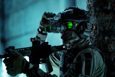 Man in uniform with machine gun and turned on night vision device inside broken building. Closeup airsoft soldier with green light on face in night building.の写真素材