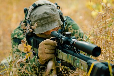 Closeup airsoft man in uniform lay in long grass with sniper rifle. Soldier aims at the sight.の写真素材