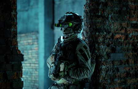 Man in uniform with machine gun and turned on night vision device stand between two brick walls. Airsoft soldier with green light on face in night building.の写真素材