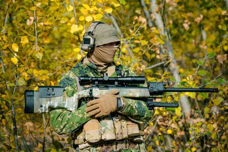 Airsoft man in uniform hold sniper rifle on yellow forest backdrop. Side view.の写真素材