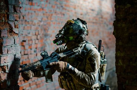 Man in uniform with machine gun and turned on night vision device move between brick wall. Closeup airsoft soldier with green light on face in night building.の写真素材