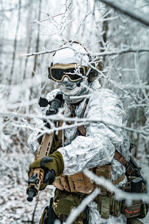 Airsoft man in white camouflage uniform and machinegun with optical sight. Soldier in the winter forest between branches. Vertical photoの写真素材