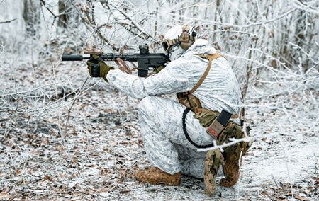Airsoft man in white camouflage uniform with machinegun. Soldier  stood on knelt in the winter forest and aims at the sight of the machinegun. Side viewの写真素材