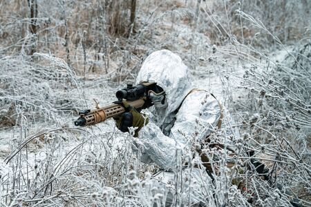 Military man in white camouflage uniform with hood and machinegun in the long winter grass. Soldier stood on knelt and aims of the machinegun.の写真素材