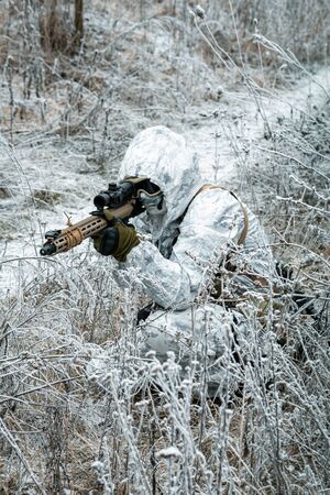 Military man in white camouflage uniform with hood and machinegun in the long winter grass. Soldier stood on knelt and aims of the machinegun. Vertical photoの写真素材