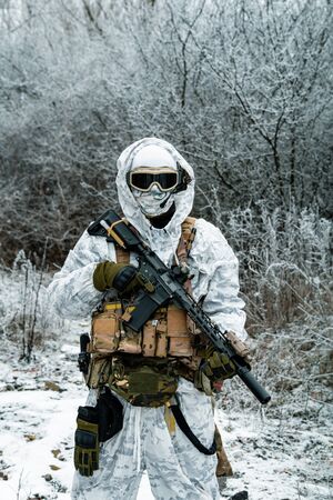 Military man in white camouflage uniform with hood and machinegun. Soldier in the winter forest territory background. Vertical photoの写真素材