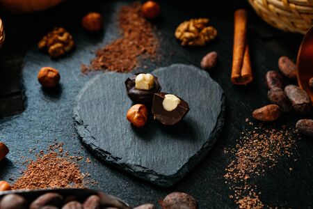 Cut handmade chocolate candy with haselnut on blackstone stand in heart shape. Chocolate candies with cocoa beans, nuts, cocoa powder around.の写真素材