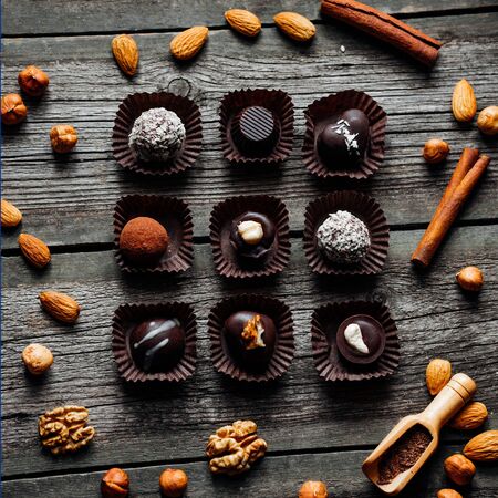 Nine handmade chocolate candies inside of candy wrappers on vintage wooden background. Nuts, cinnamon, almond, husel nut around. Top view.の写真素材