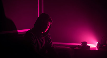 Young man in hoodie sitting on couch in dark room with neon light.の素材