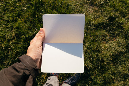 partial view of person holding book with blank pages in park during sunny dayの写真素材