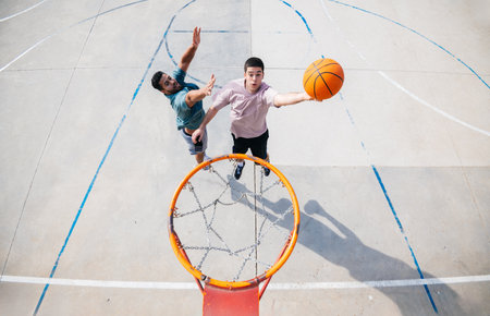 Two young men playing basketball on a sunny day, one jumping to score a basket while the other tries to block himの写真素材