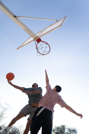 Two male friends jumping to score a basket while playing basketball outdoors on a sunny dayの写真素材