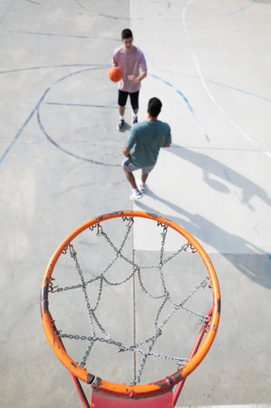 Two friends playing basketball outdoors, passing the ball on a sunny day, with the focus on the metal hoop and chain netの写真素材