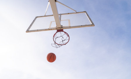 Orange basketball falling into the hoop against a blue sky backgroundの写真素材