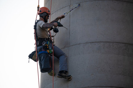 Industrial climber using a power drill while hanging on ropes to perform maintenance work on a...