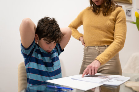 Mother assisting her son with homework, pointing at a book on the table, in a home learning environmentの写真素材