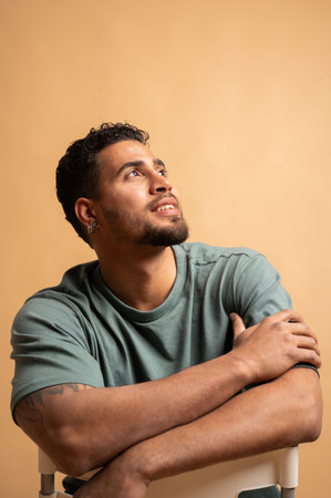 Studio portrait of a young Colombian man sitting on a chair, looking up with a hopeful and positive...