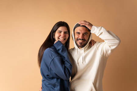 Cheerful man and woman wearing hoodies, laughing and embracing on a beige background, showing genuine happiness and connectionの写真素材