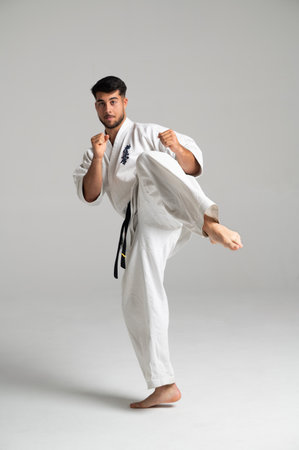 Young man practicing kyokushinkai karate, kicking high wearing a white keikogi and black belt in a bright studio environmentの写真素材