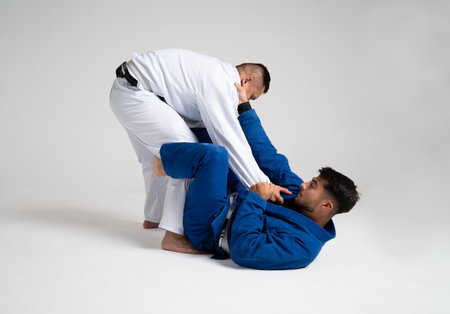 Two male athletes practicing jiu jitsu, grappling on the floor of a white studio, demonstrating a...
