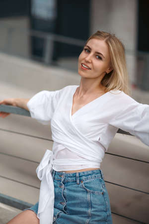 Beautiful blonde fashionable sexy tourist girl walking in city street. Woman looking at camera outdoors wearing trendy white t-shirt, denim jean short skirt.の写真素材