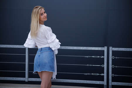 Beautiful blonde fashionable sexy tourist girl walking in city street. Woman looking at camera outdoors wearing trendy white t-shirt, denim jean short skirt.の写真素材