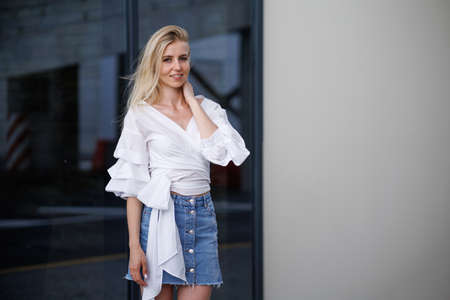 Beautiful blonde fashionable sexy tourist girl walking in city street. Woman looking at camera outdoors wearing trendy white t-shirt, denim jean short skirt.の写真素材