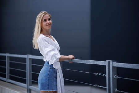 Beautiful blonde fashionable sexy tourist girl walking in city street. Woman looking at camera outdoors wearing trendy white t-shirt, denim jean short skirt.の写真素材
