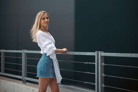 Beautiful blonde fashionable sexy tourist girl walking in city street. Woman looking at camera outdoors wearing trendy white t-shirt, denim jean short skirt.の写真素材