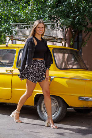 Beautiful young stylish hipster girl in fashionable summer clothes sitting on a bright yellow vintage car on the streetの写真素材