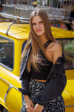 Beautiful young stylish hipster girl in fashionable summer clothes sitting on a bright yellow vintage car on the streetの写真素材