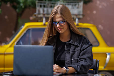Professional young business woman in sexy glasses in black jeans jacket is working on a laptop on the Internet with a new financial project. A freelance girl works remotely in a caの写真素材