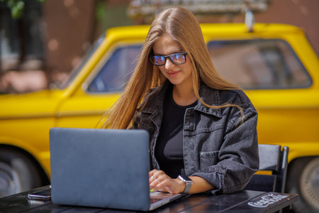 Professional young business woman in sexy glasses in black jeans jacket is working on a laptop on the Internet with a new financial project. A freelance girl works remotely in a caの写真素材