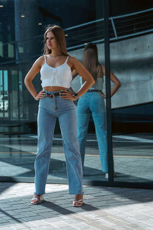 Trendy young woman in white t-shirt in vintage jeans in white shoes walks on city. Attractive girl in casual clothes is walking on the street. american style.の写真素材