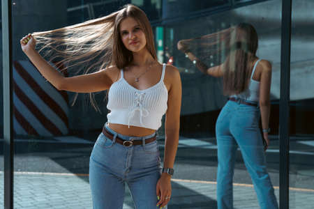 Trendy young woman in white t-shirt in vintage jeans in white shoes walks on city. Attractive girl in casual clothes is walking on the street. american style.の写真素材