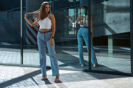 Trendy young woman in white t-shirt in vintage jeans in white shoes walks on city. Attractive girl in casual clothes is walking on the street. american style.の写真素材