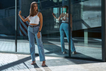 Trendy young woman in white t-shirt in vintage jeans in white shoes walks on city. Attractive girl in casual clothes is walking on the street. american style.の写真素材