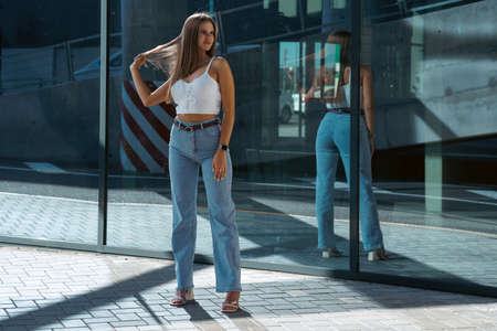 Trendy young woman in white t-shirt in vintage jeans in white shoes walks on city. Attractive girl in casual clothes is walking on the street. american style.の写真素材