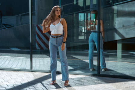 Trendy young woman in white t-shirt in vintage jeans in white shoes walks on city. Attractive girl in casual clothes is walking on the street. american style.の写真素材