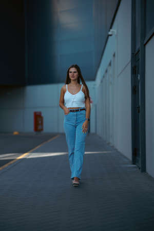 Trendy young woman in white t-shirt in vintage jeans in white shoes walks on city. Attractive girl in casual clothes is walking on the street. american style.の写真素材