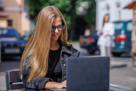 Professional young business woman in sexy glasses in black jeans jacket is working on a laptop on the Internet with a new financial project. A freelance girl works remotely in a caの写真素材
