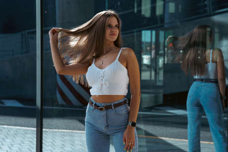 Trendy young woman in white t-shirt in vintage jeans in white shoes walks on city. Attractive girl in casual clothes is walking on the street. american style.の写真素材