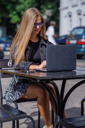 Professional young business woman in sexy glasses in black jeans jacket is working on a laptop on the Internet with a new financial project. A freelance girl works remotely in a caの写真素材